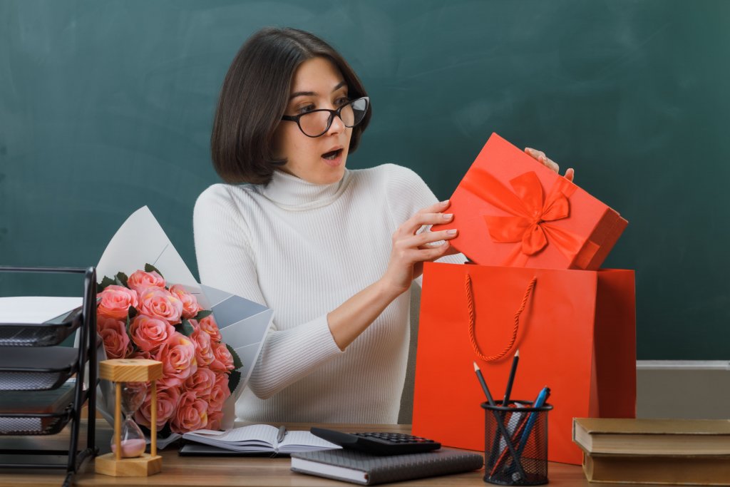 surprised-young-female-teacher-holding-and-looking-at-present-sitting-at-desk-with-school-tools-in-classroom (1).jpg